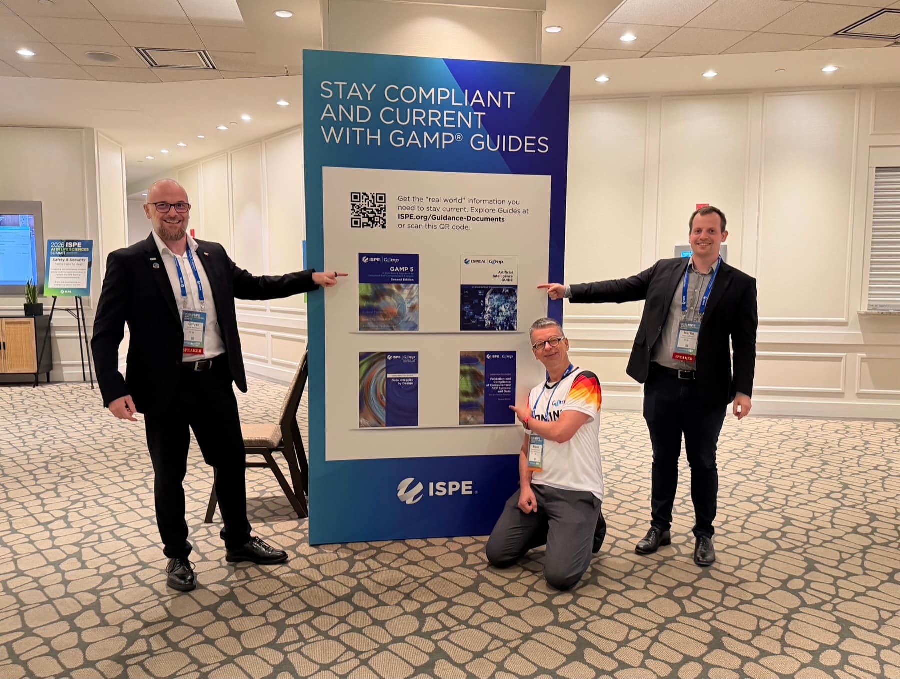 Frank Henrichmann, Oliver Herrmann und Martin Heitmann zeigen am ISPE-Stand des AI in Life Sciences Summit 2026 auf ausgestellte GAMP-Leitf&auml;den, darunter GAMP 5